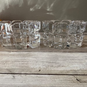 May include: Two clear glass candle holders with a geometric design. The holders are made of thick glass and have a square shape.