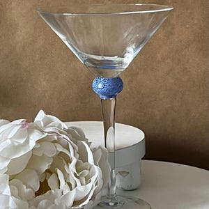 Peut inclure: Un verre à martini transparent avec une boule bleue texturée sur la tige. Une fleur blanche est au premier plan. Le verre est sur une surface blanche avec un fond marron.