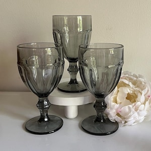 May include: Three gray glass goblets with a textured design. The goblets are on a white surface with a white flower in the background.