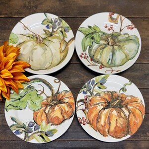 May include: Four white ceramic plates with hand-painted pumpkin designs. Each plate features a different pumpkin, with green leaves and berries. The pumpkins are in shades of orange, green, and white, with brown stems. The plates are arranged on a dark wood surface.