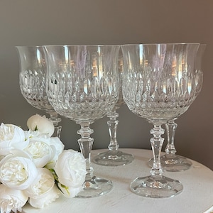 Puede incluir: Cuatro copas de vino de cristal transparente con intrincados patrones tallados. Las copas tienen forma de tulip&aacute;n y est&aacute;n sobre una superficie blanca. Flores blancas en primer plano. Las copas son elegantes y adecuadas para ocasiones formales.