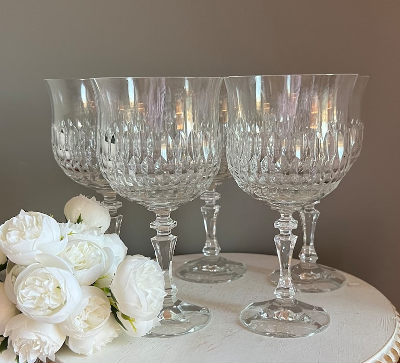 Puede incluir: Cuatro copas de vino de cristal transparente con intrincados patrones tallados. Las copas tienen forma de tulip&aacute;n y est&aacute;n sobre una superficie blanca. Flores blancas en primer plano. Las copas son elegantes y adecuadas para ocasiones formales.