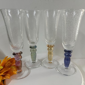 May include: Four clear champagne flutes with textured glass. Each flute has a different colored stem: purple, green, yellow, and blue. The flutes are on a white surface, with an orange flower in the foreground.