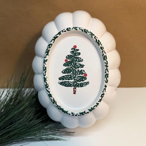 May include: White, scalloped-edge Christmas plate with a green and red Christmas tree design. The plate has a green and white patterned border. The plate is a festive holiday decoration.