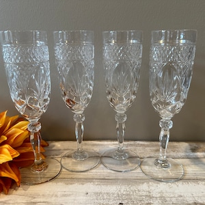 May include: Four clear glass champagne flutes with a cut crystal design. The flutes are arranged in a row on a wooden surface.