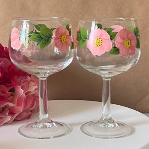 May include: Two clear wine glasses decorated with pink floral patterns and green leaves. The flowers have yellow centres. The stemware is on a white surface with a pink flower arrangement in the background.
