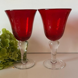 May include: Two red glass wine goblets with clear glass stems and bases. The goblets have a rounded bowl shape.