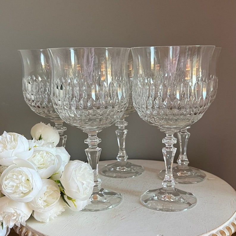 Puede incluir: Cuatro copas de vino de cristal transparente con intrincados patrones tallados. Las copas tienen un cuenco en forma de tulip&aacute;n, un tallo delgado y una base redonda. Flores blancas en primer plano a&ntilde;aden elegancia.