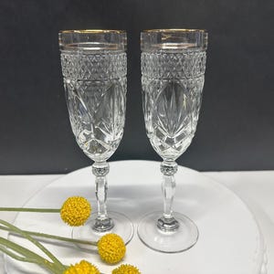 May include: Two clear crystal champagne flutes with gold rims. The glasses have a diamond-cut pattern and sit on a clear stem with a decorative base. Yellow flowers are in the foreground.