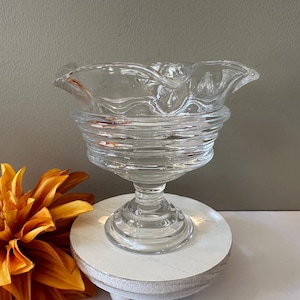 May include: A clear glass bowl with a ruffled edge and a pedestal base. The bowl has a fluted design and is perfect for serving desserts or snacks.