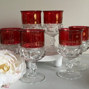 May include: Set of six red and clear glass stemmed drinking glasses with a textured pattern. The glasses have a ruby red colored base and a clear glass stem and bowl.