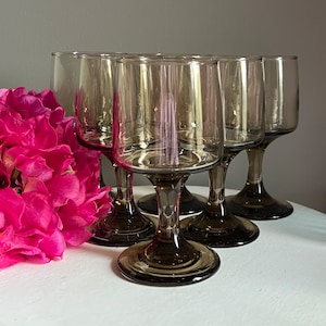 May include: Six vintage-style wine glasses in a smoky grey hue. The glasses feature a square bowl design and a rounded base. They are arranged on a white surface, next to a cluster of vibrant pink flowers.