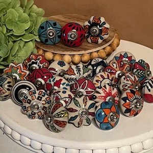 May include: A collection of round ceramic cabinet knobs featuring various hand-painted floral and geometric patterns in vibrant colors including red, blue, orange, green, and black, each with a central silver-toned metal screw and hardware.