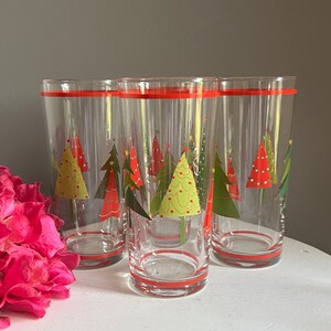 May include: Set of four clear glass drinking glasses with a red band around the top. Each glass features a festive design of green, red and yellow Christmas trees with white accents.