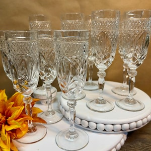May include: A set of clear crystal champagne flutes with detailed diamond and floral designs. The glasses are displayed on a white beaded tray, with a yellow and orange flower in the foreground. The stemware is vintage and elegant.