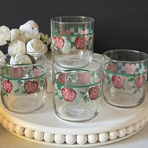 May include: Set of four clear drinking glasses. Each glass features a repeating pattern of red apples, green leaves, and a green and light blue checkered border. The glasses are displayed on a white, round, beaded tray.