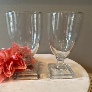 May include: Two clear glass goblets with square bases. The goblets are empty and sitting on a white surface.