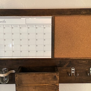 May include: A brown wooden organiser with a white dry erase calendar, a cork board, a small wooden box, a glass jar with a leather strap, and two metal hooks.