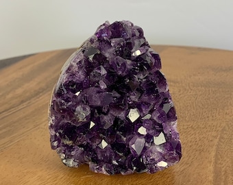 May include: A large amethyst crystal cluster with a deep purple color. The crystal is made up of many smaller crystals that are clustered together. The crystal is on a wooden surface.
