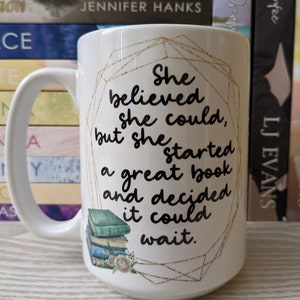 May include: White ceramic mug with a gold geometric design and a black quote that reads "She believed she could, but she started a great book and decided it could wait." The quote is surrounded by a stack of books with floral accents.