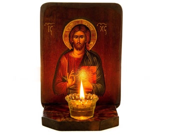 Op de afbeelding: Een houten icoon van Jezus Christus met een brandende kaars ervoor. Het icoon is geschilderd in een traditionele stijl en heeft een rode achtergrond met gouden accenten. De kaars staat in een transparante glazen houder en brandt helder.