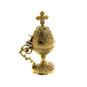 May include: A gold-coloured censer with detailed floral and scrollwork patterns. It has a cross on top and a decorative handle. The base is a pedestal design. The censer is likely used in religious ceremonies.