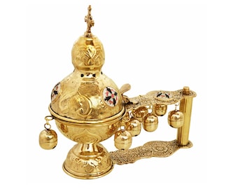 May include: Ornate gold-colored censer with intricate floral and geometric patterns. The censer features a bulbous body, a decorative handle, and small bells. The design includes small, colorful, circular accents. A cross tops the lid.