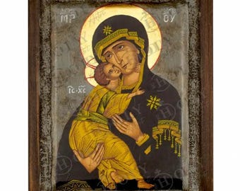 May include: A framed religious icon depicting a woman holding a child. The woman wears a black robe and a gold halo. The child is dressed in yellow. The icon has a dark wooden frame and is set against a textured background.