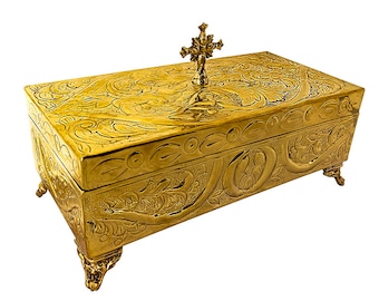 May include: A gold-colored rectangular box with ornate floral and scrollwork carvings. The box has a decorative cross on the lid and rests on four ornate feet. The box is made of metal and has a polished finish.