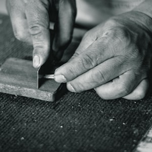 Puede incluir: Se muestran las manos de una persona afilando una herramienta de metal sobre un trozo de madera. La madera tiene una textura rugosa y est&aacute; marcada con n&uacute;meros.