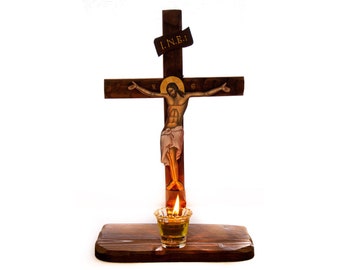 May include: A wooden crucifix with a depiction of Jesus Christ on the cross. The crucifix is mounted on a wooden base with a small glass candle holder containing a lit candle. The inscription "I.N.R.I." is visible on the cross.