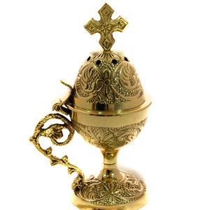 May include: A gold-colored censer with intricate floral and scrollwork designs. The censer features a cross on top and a decorative handle. The base is round and the censer is elevated on a pedestal.