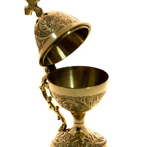 May include: A gold-colored censer with an open lid and ornate floral and scrollwork patterns. The censer has a handle and a cross-shaped finial on top. The chalice-shaped bowl sits on a decorative base.