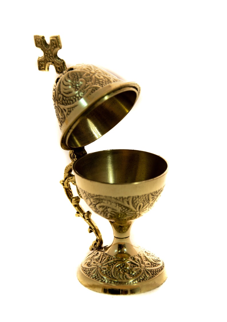 May include: A gold-colored censer with an open lid and ornate floral and scrollwork patterns. The censer has a handle and a cross-shaped finial on top. The chalice-shaped bowl sits on a decorative base.