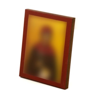 May include: A framed icon with a blurred image of a person in a red and yellow robe. The frame is made of wood with a red border.