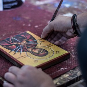 May include: A person is working on a framed religious icon with a gold background. The icon depicts a woman with multiple swords piercing her body. The text on the icon reads "HOLY MARTYR" and "AGAPIA".