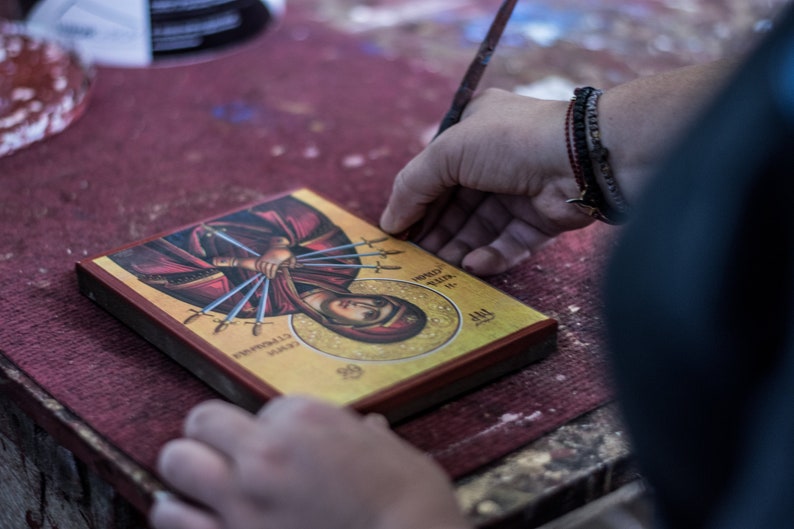 May include: A person is working on a framed religious icon with a gold background. The icon depicts a woman with multiple swords piercing her body. The text on the icon reads "HOLY MARTYR" and "AGAPIA".