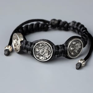 Op de afbeelding: Een zwarte koordarmband met drie ronde, zilverkleurige metalen bedels met reliëf van religieuze figuren. De armband is gemaakt met een geknoopt vlechtpatroon en heeft twee verstelbare uiteinden afgewerkt met kleine zilverkleurige metalen kralen.