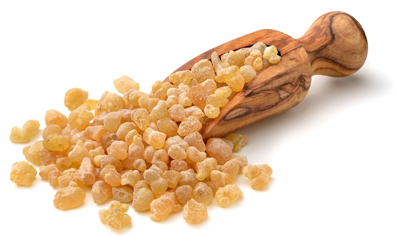 May include: A wooden scoop filled with golden brown frankincense resin, spilling out onto a white surface.
