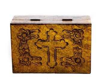 May include: A wooden box with a hinged lid. The front of the box features a carved cross in the center, flanked by floral designs. The wood has a warm, golden-brown hue.