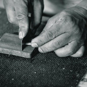 May include: A person's hands are shown sharpening a metal tool on a piece of wood. The wood has a rough texture and is marked with numbers.