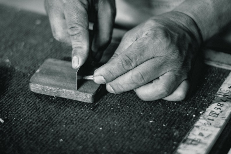May include: A person's hands are shown sharpening a metal tool on a piece of wood. The wood has a rough texture and is marked with numbers.