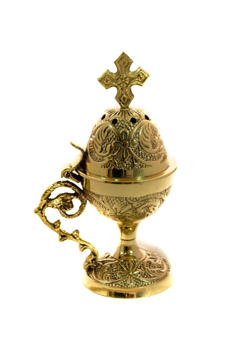 May include: A gold-colored censer with intricate floral and scrollwork designs. The censer features a cross on top and a decorative handle. The base is round and the censer is elevated on a pedestal.