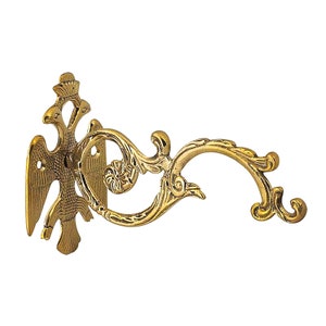 Puede incluir: Soporte de pared de latón dorado ornamentado con un diseño de águila. El soporte presenta un brazo curvo con detalles florales y de volutas. La pieza decorativa está diseñada para sostener un objeto colgante y tiene una estética vintage.
