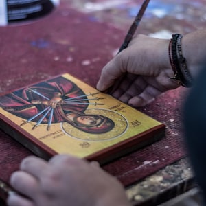 May include: A person is working on a framed religious icon with a gold background. The icon depicts a woman with multiple swords piercing her body. The text on the icon reads "HOLY MARTYR" and "AGNES".
