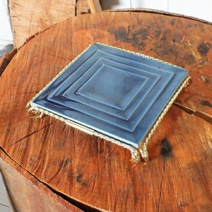 Könnte beinhalten: Eine blaue quadratische Keramikfliese mit einem goldenen Metallrahmen und dekorativen Beinen. Die Fliese hat ein konzentrisches quadratisches Design.