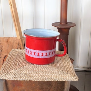 Peut inclure: Pichet en émail rouge avec un bord blanc et un motif floral blanc. Le pichet a une poignée et est posé sur un dessous de plat en osier.