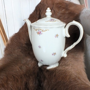 Könnte beinhalten: Eine weiße Porzellan-Kaffeekanne mit Blumenmuster und goldenen Akzenten. Die Kanne hat einen Deckel und einen Griff. Sie steht auf einem braunen Fellteppich.