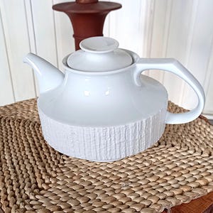 Könnte beinhalten: Eine weiße Keramik-Teekanne mit einem strukturierten, rindenartigen Design auf der unteren Hälfte. Die Teekanne hat einen gebogenen Griff und einen Deckel mit einem kleinen Knopf. Sie steht auf einer gewebten Matte.