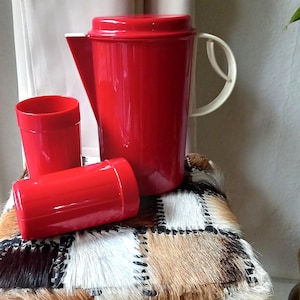 Könnte beinhalten: Ein roter Kunststoffkrug mit weißem Griff und Deckel, zusammen mit zwei roten Bechern. Der Krug ist hoch und zylindrisch, mit einem Ausguss zum Ausgießen. Die Becher sind ebenfalls rot und zylindrisch. Die Artikel werden auf einem Patchwork-Kissen präsentiert.